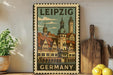 A vintage-style metal sign with a colorful illustration of a German city, featuring a clock tower and other architectural elements, is displayed on a shelf next to a wooden cutting board and a potted plant.