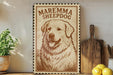 a framed illustration of a large, fluffy dog with a brown and white coat, sitting in a grassy field. The dog appears to be a breed called Maremma Sheepdog, as indicated by the text on the frame. The frame is placed on a shelf next to a wooden cutting board and a potted plant, creating a cozy and homey atmosphere.