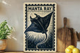 a framed print of a manta ray, a type of ray, on a wall. The manta ray is depicted in a stylized, black and white illustration with a decorative border. The print is placed next to a wooden cutting board and a potted plant, creating a homely, coastal-inspired setting.