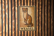 A weasel illustration on a wooden panel with the word "WEASEL" below it.