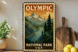 A vintage-style metal sign with a mountain landscape and pine trees, featuring the text "OLYMPIC NATIONAL PARK USA".