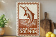 a sign with a dolphin illustration, placed on a shelf next to a wooden cutting board and some lemons.