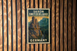 A vintage postage stamp featuring a scenic mountain landscape with the words "SAXON SWITZERLAND" and "GERMANY" printed on it, hanging on a wooden wall.