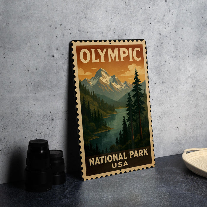 A vintage-style metal stamp depicting a scenic landscape with mountains, pine trees, and a river, with the text "OLYMPIC NATIONAL PARK USA" printed on it.