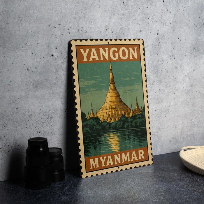 A vintage-style metal sign with the text "YANGON MYANMAR" and an illustration of a Buddhist temple, placed on a gray surface next to some black camera lenses.