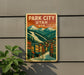 A vintage-style metal sign with the text "PARK CITY UTAH USA" and an illustration of a mountain town with colorful buildings and a winding road.