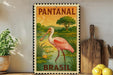 A vintage-style stamp depicting a pink flamingo standing in a pond, with the text "PANTANAL BRASIL" on it. The stamp is displayed on a shelf next to a wooden cutting board and a potted plant.