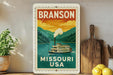 A vintage-style metal sign with the text "BRANSON MISSOURI USA" and an illustration of a steamboat on a river, mounted on a wall next to a wooden cutting board and a potted plant.