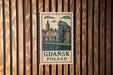 A vintage-style sign with a drawing of a cityscape and the text "Gdansk Poland" is displayed on a wooden wall.