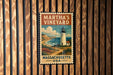 A vintage-style metal sign with a lighthouse and coastal landscape, mounted on a wooden wall.