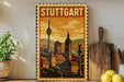 A vintage-style metal sign with the text "STUTTGART" displayed prominently, featuring a cityscape illustration with a tower and other architectural elements. The sign is placed on a wooden surface, with a potted plant, a wooden cutting board, and some lemons nearby.