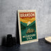 A vintage-style metal sign with the text "BRANSON MISSOURI USA" and an illustration of a steamboat on a river, placed on a gray countertop next to some black camera lenses.