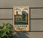 A sign with a picture of a town and the text "Gdansk Poland" is displayed on a wall, with a plant to the left of the sign.