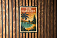 A vintage-style poster of New Caledonia, a country in the South Pacific, featuring a tropical beach scene with palm trees and a sunset over the ocean.