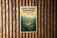 A vintage-style postage stamp featuring a scenic mountain landscape with pine trees and a sun in the sky, mounted on a wooden wall.