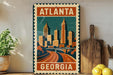 A vintage-style metal sign with the text "ATLANTA GEORGIA" and an illustration of a city skyline, placed on a wooden surface next to a wooden cutting board and some potted plants.