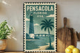 A vintage-style metal sign depicting a lighthouse and a palm tree, with the text "PENSACOLA FLORIDA USA" on it, is displayed on a shelf next to a wooden cutting board and some potted plants.
