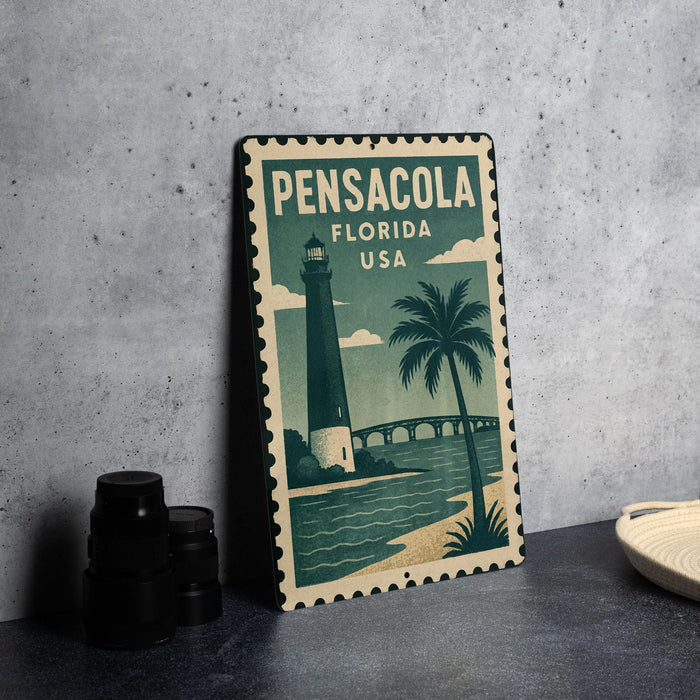 A vintage-style metal sign with the text "PENSACOLA FLORIDA USA" and an illustration of a lighthouse and palm tree, placed on a gray countertop next to some black camera lenses.