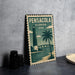 A vintage-style metal sign with the text "PENSACOLA FLORIDA USA" and an illustration of a lighthouse and palm tree, placed on a gray countertop next to some black camera lenses.