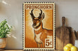 A framed stamp depicting a Pronghorn antelope, with the text "PRONGHORN" and the number "5c" printed on it. The stamp is placed on a shelf next to a wooden cutting board and a potted plant.