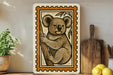a framed illustration of a koala bear on a tree branch, placed on a shelf next to a wooden cutting board and a potted plant.