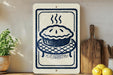 a kitchen scene with a pie on a wooden board, a potted plant, and a wooden cutting board.