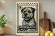 A black and white illustration of a dog, likely a breed from the Netherlands, is displayed on a white postcard-like frame. The frame is placed on a wooden surface, with a potted plant, a wooden cutting board, and some lemons nearby.