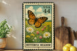 A framed postage stamp depicting a butterfly in a meadow, with the text "Butterfly Meadow" and the number "44" printed on it. The stamp is placed on a shelf next to a wooden cutting board and a potted plant.