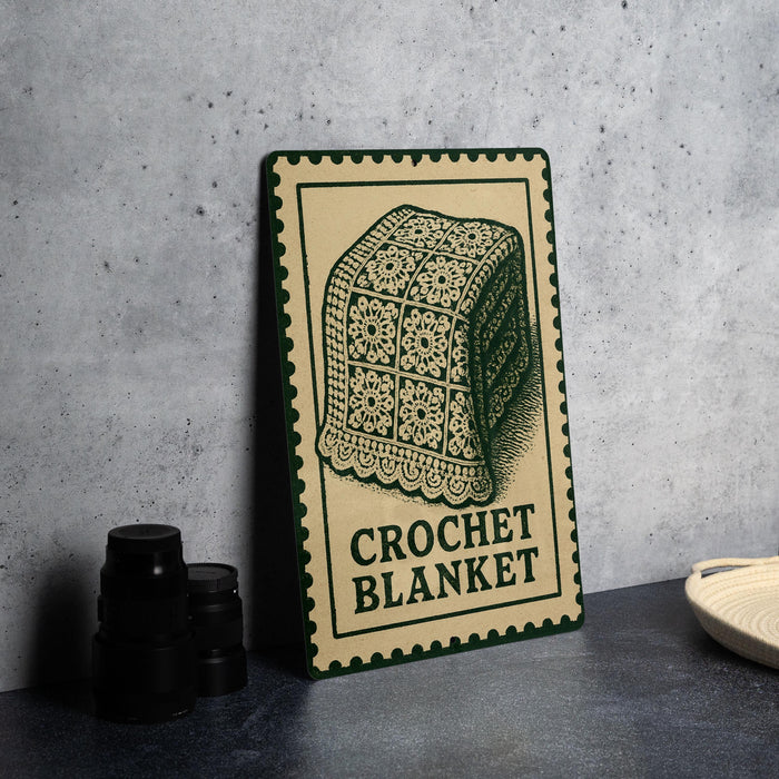 A framed image of a crocheted blanket is displayed on a table, accompanied by a stack of black camera lenses and a white dish.