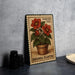 A metal sign with a floral design, featuring a potted plant with red flowers, is displayed on a table.