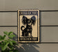A metal sign with a black and white illustration of a Russian toy dog, mounted on a wall next to a plant.