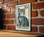 A metal sign featuring a cat illustration is mounted on a brick wall.