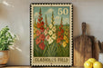 A framed stamp depicting a field of colorful flowers, with the text "GLADIOLUS FIELD" printed on it. The stamp is placed on a shelf next to a wooden cutting board and a potted plant.
