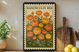 a framed illustration of a flower with orange petals, surrounded by green leaves, placed on a shelf next to a wooden cutting board and a potted plant.