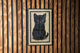A black cat illustration is displayed on a wooden wall, framed within a decorative border.