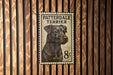 A metal postage stamp featuring a black dog, specifically a patterdale terrier, is displayed on a wooden wall.