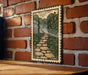 A framed illustration of a stone path leading through a lush, green landscape is displayed on a brick wall.