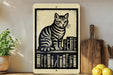 A framed image of a striped cat sitting on a bookshelf, surrounded by books, is displayed on a shelf next to a wooden cutting board and a potted plant.
