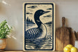 a wooden sign with a black and white illustration of a loon swimming in a lake, surrounded by trees and a cloudy sky.