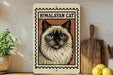 A framed image of a Himalayan cat is displayed on a shelf, accompanied by a wooden cutting board, a potted plant, and some lemons.