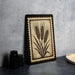 a framed artwork depicting a wheat stalk with three ears, placed on a dark surface next to a stack of black camera lenses.