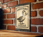 A metal sign with a black and white illustration of a goose and the text "Barnyard Goose" is mounted on a brick wall.