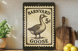 A black and white postage stamp featuring a goose, with the text "BARNYARD 5c GOOSE" printed on it. The stamp is placed on a shelf next to a wooden cutting board and some kitchen utensils.