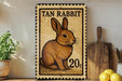 A tan rabbit on a postage stamp, with the text "TAN RABBIT" and "20c" printed on it. The stamp is placed on a wooden surface, with a potted plant, a wooden cutting board, and some lemons nearby.