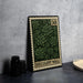 A framed print of a postage stamp with a green floral design, placed on a table next to some black camera lenses.