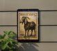 a black and white illustration of a goat on a wall, with a postage stamp-like border.