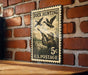 A vintage U.S. postage stamp featuring a duck hunting scene, mounted on a brick wall.