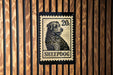 A black and white stamp-like design featuring a dog's head and the word "SHEPDOG" printed on a wooden background.