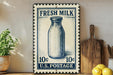 A vintage-style stamp depicting a glass milk bottle, with the text "FRESH MILK" and "10¢ U.S. POSTAGE" printed on it. The stamp is placed on a shelf next to a wooden cutting board and a potted plant.
