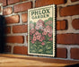 A vintage-style stamp with a floral illustration is displayed on a brick wall, featuring the text "PHLOX GARDEN".